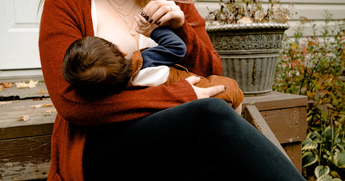 Mamma i rost-cardigan över vit topp ammar barn utomhus, lager-på-lager-teknik