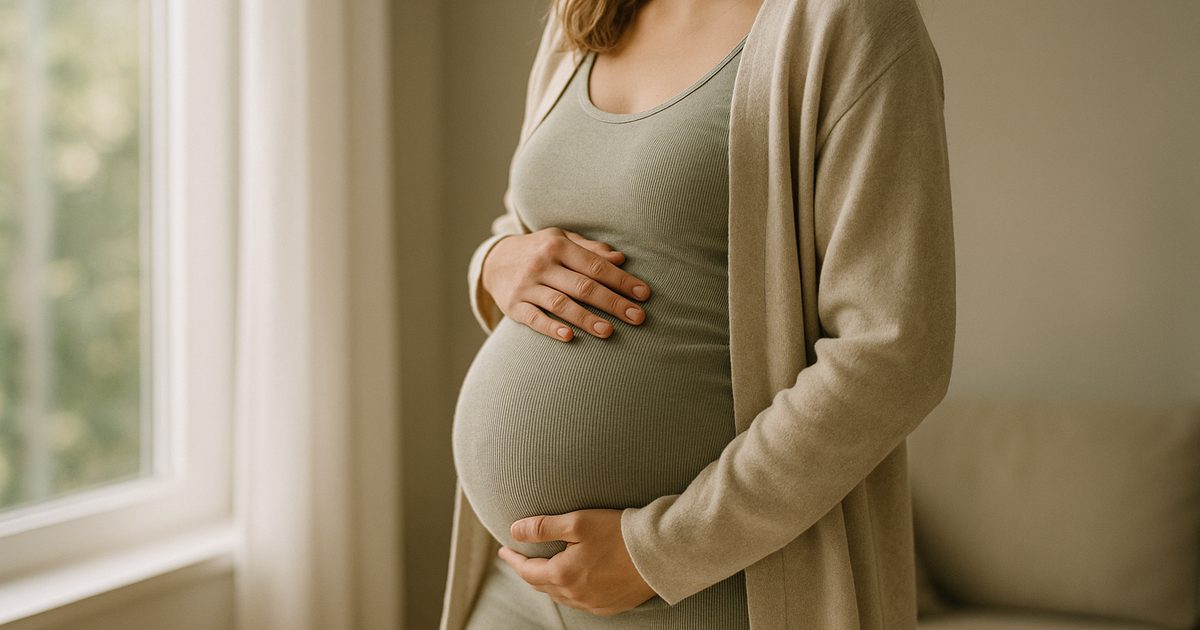 Gravid kvinna i mjuk neutral hemklädsel vid ett soligt fönster, sett från sidan utan ansikte, med handen vilande mot magen