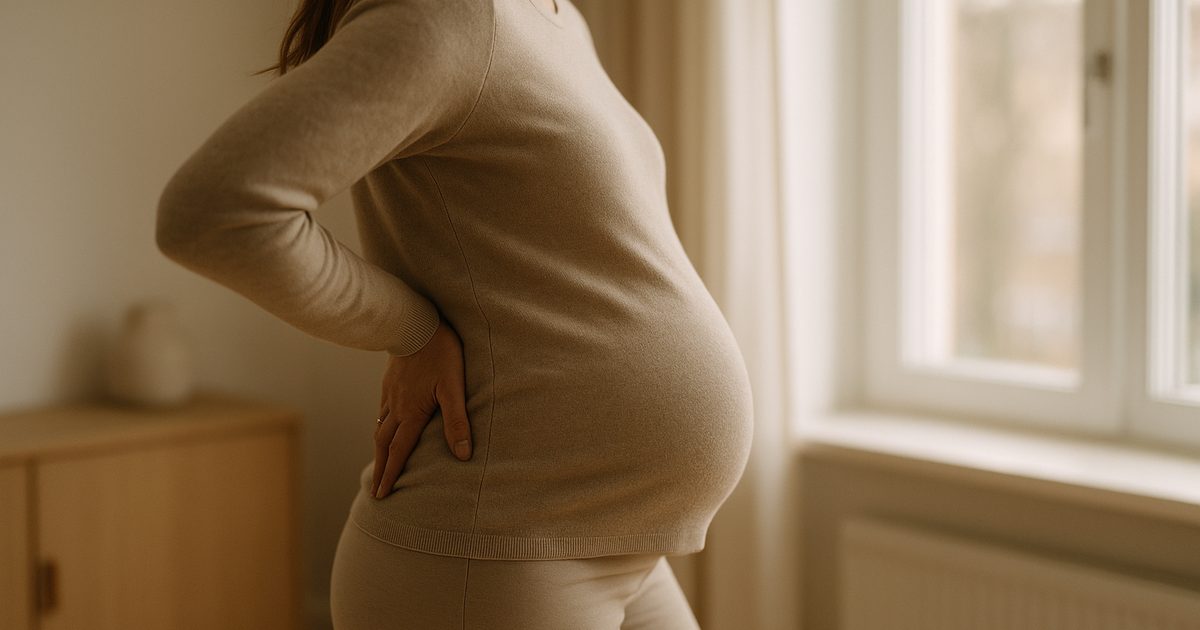 Gravid kvinna sedd bakifrån i mjuk klädsel vid ett fönster, ena handen vilande mot nedre ryggen, lugnt ljus