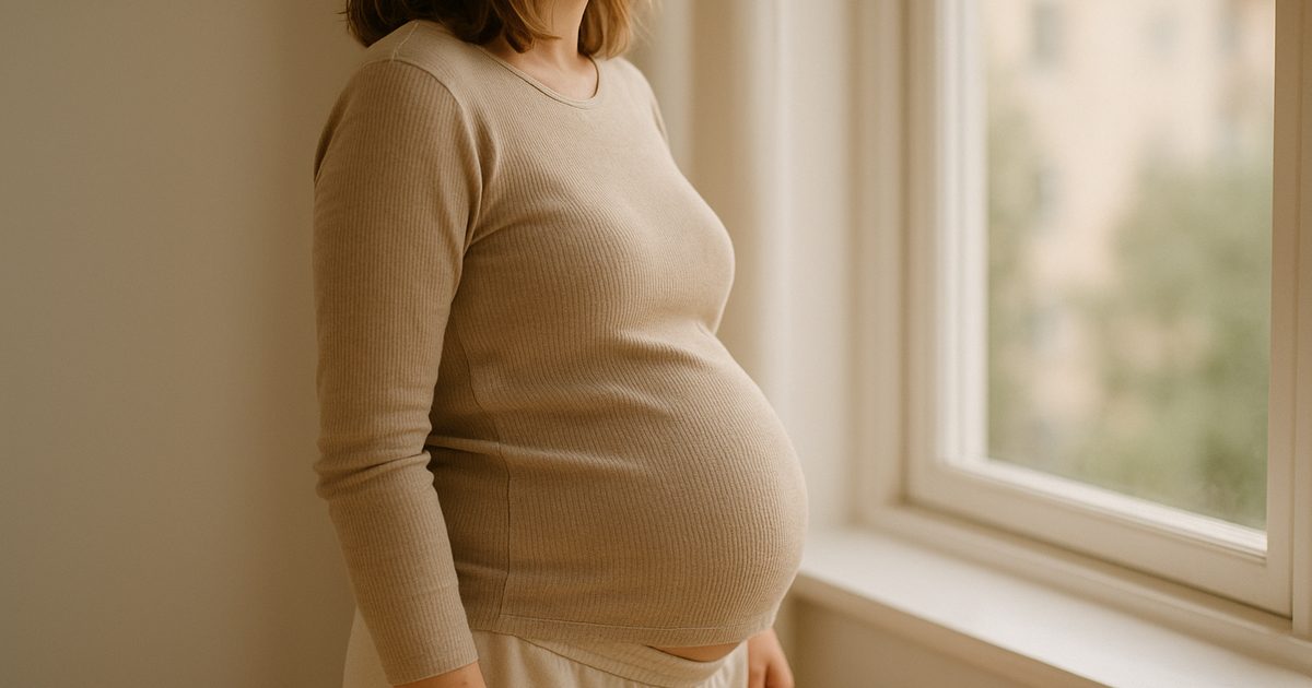 Gravid kvinna står avslappnad vid ett fönster utan bälte, sedd från sidan utan ansikte