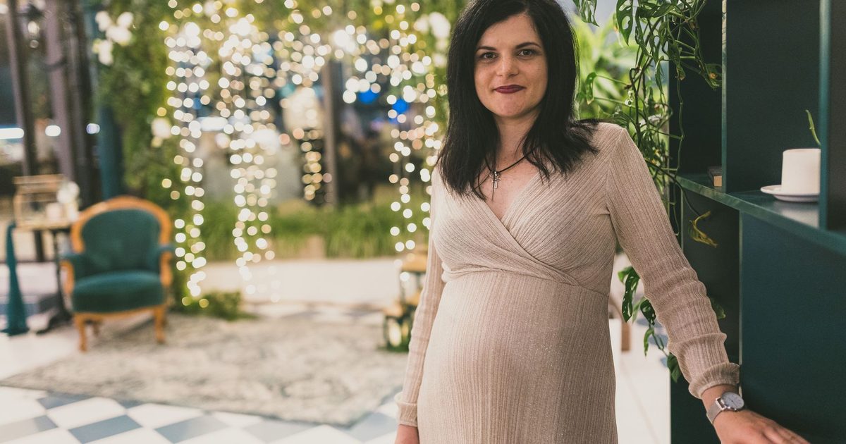 Pregnant woman in elegant dress standing indoors with soft lighting and greenery.