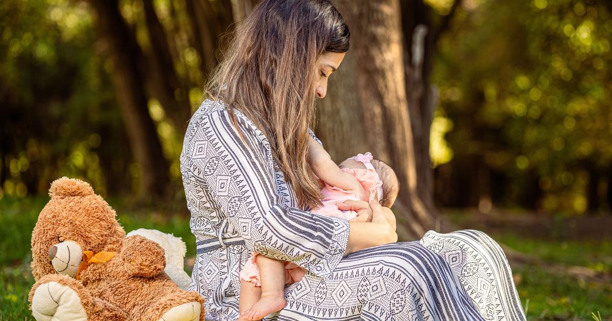 Tender moment of a mother breastfeeding her baby outdoors with a teddy bear nearby.